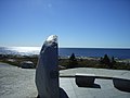 Photo d'une pierre demi-circulaire vue depuis sa tranche, avec l'océan et le ciel bleu en arrière-plan. De petits bancs de pierre se trouvent près du mémorial, à droite de l'image.