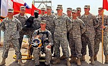 Thirteen US Army soldiers are posing for another camera (not seen) with a statuette of their beaver mascot and a red guidon. One soldier is sitting in front of the rest, wearing a harness and yellow diver's helmet. To the back-right of the group, a number of flags wave in the wind.