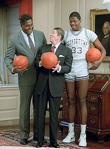 Two tall African-American men, one in a suit, one in a gray basketball uniform, stand behind a shorter elderly white male in an ornate room, with each man holding a basketball.