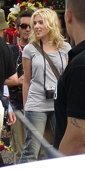 A young woman with tousled medium length blonde hair loosely around her shoulders and face, looking to her right, her right arm slightly behind her, stands in the middle of a group of men. She is dressed in denim jeans, a light gray short sleeved shirt and a camera hangs around her neck.