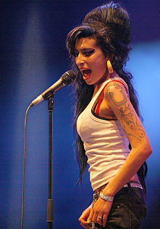 Amy Winehouse performing on 29 June 2007 at the Eurockéennes music festival in eastern France. Winehouse is looking into the crowd while singing sidelong into a handheld microphone. Her black hair is combed back in a relaxed style. Her tattooed shoulder is bare and she is wearing large, chunky earrings.