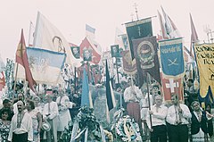 Several people wearing the traditional Ukrainian clothing, vyshyvanka, carry a variety of regional flags and portraits of national leaders as they stand around a headstone adorned with Ukrainian national symbolism.