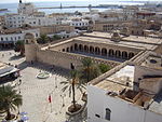 An aerial view of a town square. The inside of a large, square building is visible.