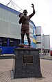 A statute of a man holding a trophy and lifting his left arm into the air. The statute is in front of a football stadium