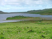 A small island in a lake lies offshore from green fields. A small wooden footbridge leads to the islet which contains various stone ruins including at least two gable ends.