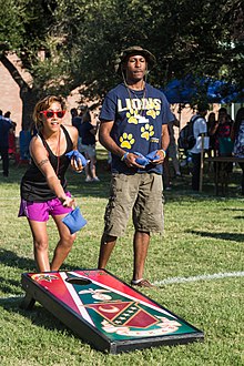 A woman tossing a cornhole bag in front of her board while a man stands beside her