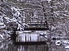 Snow covers a simple wooden footbridge which crosses a stream in a forest