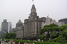 Pedestrians walk before a row of trees and a series of tall buildings. A blue sky overhead is obscured slightly by several clouds.