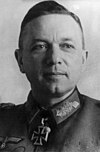 The head of a man, shown in semi-profile. He wears a military uniform with a military decoration in shape of an iron cross displayed at the front of his shirt collar. His hair is dark and combed to back.