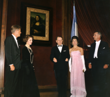 Three men and two women stand near the Mona Lisa. All are dressed formally, one woman in a spectacular pink gown.