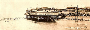 A black-and-white postcard shows a photograph, taken from a location on the water, of a large building sitting on pier by the beach. The beach is fronted by a seawall and a crowded waterfront beyond. The caption on the postcard says, "Where life is worth living at Galveston".
