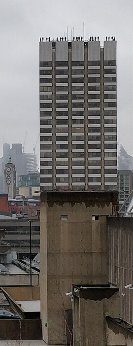 A photograph of the Project84 installation in London. A tall tower block - at the apex, statues of men stand disturbingly close to the edge.