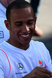 Head and shoulders of a man in his twenties, who has close-cropped black hair and is showing his teeth as he smiles while signing something unseen. He is wearing a white, V-neck T-shirt displaying the Mercedes-Benz and Vodafone logos.