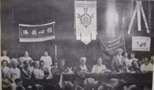 A black and white photograph depicting the head table at a women's conference surrounded by seated delegates above whom are suspended flags of various nations