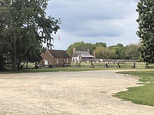 A large area of sandy dirt surrounded by grass. Behind it is a wooden rail fence enclosing a grassy area with a small brick building in the foreground and a small white shed near the back. Behind the shed is a larger wooden house longer than it is tall, painted red, with a slanted roof and two large chimneys towering over each side of the house.