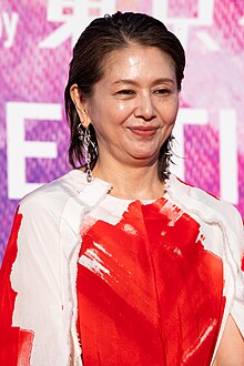 waist up portrait of Koizumi in red and white top and large silver earrings, looking to right of audience