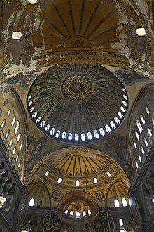 Vertical interior image of the long vaulted ceiling of the nave of Hagia Sophia showing the central ribbed dome with a ring of windows at its base, four pendentives between the four large arches supporting that main dome, two large semi-domes filling the near and far arches (with the other two arches being filled by flat walls with windows, and smaller niche semi-domes in the far large semi-dome