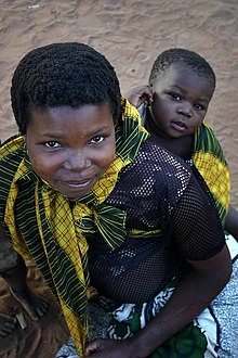 Une jeune mère sourit à l'appareil photo. À son côté se tient son bébé regardant l'appareil avec une expression de vif intérêt.