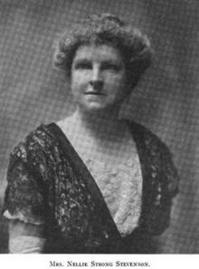 An older white woman, her grey hair in a bouffant updo, wearing a black and white lace dress.