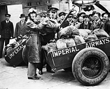 Photographie noire et blanc de Kingsford Smith tenant un sac de courrier, des chariots remplis d'autres sacs derrière.