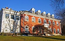 A wide brick building with dormer windows projecting from its roof and a white wooden wing on the left, seen from slightly downhill