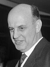 A black and white photo of a white man with thinning hair, wearing a suit and smiling.