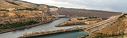 Two large channels of water flow slowly from a low sloping dam with several pipes at one end of the dam