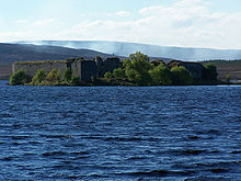 In the foreground are the blue waters of a lake. Beyond that is a wooded shoreline on which there are the ruins of a large walled structure. Smoke drifts across the moorland in the distance.