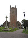 Reddish stone building with square tower. In the foreground are a cross and lamppost.