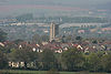 Roofs of several houses with the square tower of the church prominent amongst them. In the background are fields and hills.