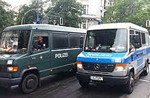 Frontale Farbfotografie von zwei Einsatzwagen der Polizei mit Fahrern. Der linke Wagen hat eine dunkelgrüne Farbe und der rechte Wagen eine blau-silber gestreifte Farbe.