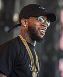 A black man, wearing glasses, a black shirt, gold chains, and a black cap, looks to his right.