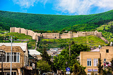 Photograph of an ochre-coloured medieval fortress on a green hilltop, with higher green hills in the background