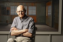 Portrait photo of artist Christopher Pratt standing with his arms folded in front of a his painting of the interior of a ruined building at Fort McAndrew, an abandoned American military base in Newfoundland.