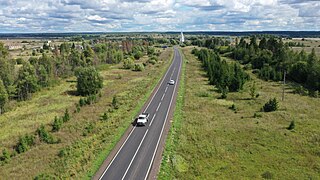 Photographie d'une route traversant un paysage mixte de champs et bosquets, avec un village possédant une église en arrière-plan.