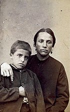 Portrait en photographie sépia d'une femme habillée simplement, au visage rehaussé de deux boucles d'oreille, et posant son bras droit sur l'épaule de son fils, au regard hagard.