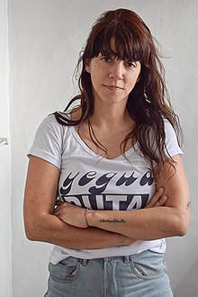 A standing woman with long brown hair, arms crossed, wearing a white T-shirt and jeans.