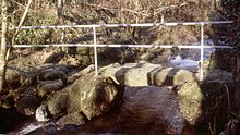 A handrail leading along a rocky creek crossing Cadnant Clapper Bridge.jpg