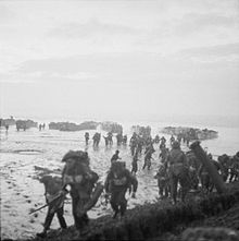 Men in the foreground burdened with equipment, behind them are mud or sand flats and in the distance landing craft