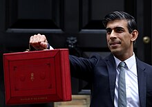 Sunak in a dark blue suit standing in front of the black door of 11 Downing Street, holding a small red suitcase at arm's length.