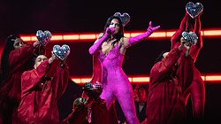 Dua Lipa performing in a pink catsuit surrounded by dancers in red outfits holding disco ball-styled hearts