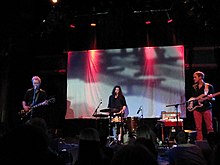 Three people performing on a dark stage illuminated by red ceiling lights. In the background, a video is projected onto a wall.