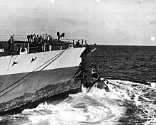 The bow of the ship, with several men standing aboard. A smaller warship is visible in the distance.