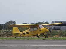 A yellow light aircraft similar to the piper j3 cub in size and design is show taxiing at a small aerodrome in Dunkeswell, England. It shares many similarities with the j3 cub