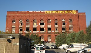 The large south-west facing side of the building, with graffiti along the top, several broken windows, and utility vans in a parking lot in the foreground