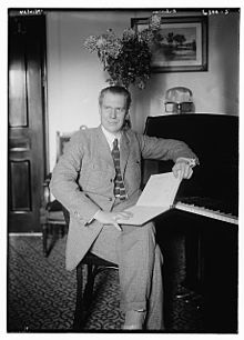 Portrait en noir et blanc d'un homme assis en costume, un livre sur ses genoux, appuyé contre un piano