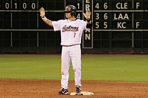 A man in a white, pinstriped baseball uniform with "ASTROS 7" on the chest and a black batting helmet stands on a base with both hands raised.