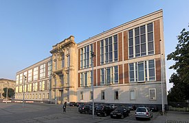 Frontale Farbfotografie von einem dreigeschossigen modernen Gebäude mit einem klassischen Mittelteil aus Steinverzierungen. An den Fenstern sind seitlich braune Steinplatten angebracht und die Fenster des oberen Stockwerks sind größer.