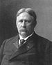 Head and shoulders of a white man with hair parted in the center and a mustache, wearing a dark suit and tie.