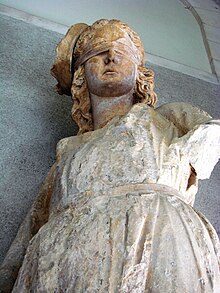 Photographie en contre-plongée d'une statue peinte endommagée et décolorée d'une figure féminine aux yeux bandés.